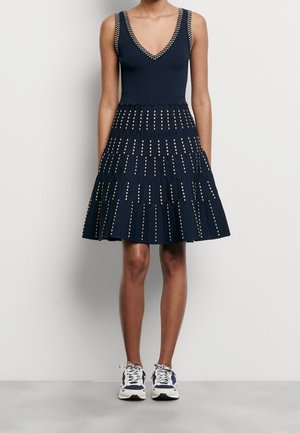 Ärmelloses marineblaues Kleid mit V-Ausschnitt und silbernen quadratischen Mustern von der Taille bis zum Saum, getragen mit weißen und marineblauen Sneakers vor einem einfarbigen Hintergrund.