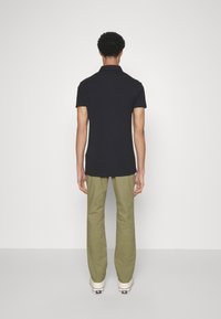 Man die met zijn rug naar de camera staat, gekleed in een zwart polo met korte mouwen, olijfgroene broek en witte sneakers, tegen een effen witte achtergrond.