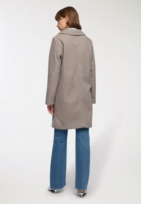 Una donna che indossa un lungo cappotto beige, jeans blu e tacchi bianchi, in piedi di spalle di fronte a uno sfondo chiaro e semplice.