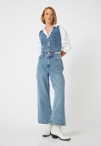 Chaleco de mezclilla con cierres de botón, combinado con jeans de pierna ancha de cintura alta en azul claro. Camisa blanca con cuello debajo. Botines blancos.