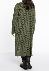 Lång olivgrön kofta med ett texturerat kabelstickat mönster. Har ribbade ärmslut och en lång silhuett, bärs över svarta byxor och stövlar.