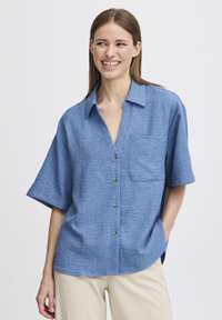 Camicia azzurro chiaro a maniche corte con bottoni, realizzata in un tessuto texturizzato, dotata di taschino sul petto e sette bottoni sul davanti.
