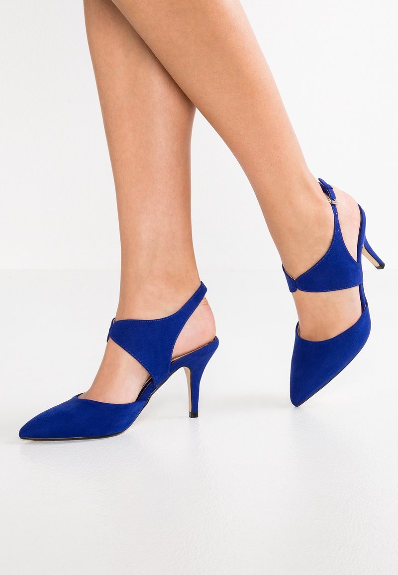 Jambes portant des escarpins slingback en daim bleu à bout pointu, avec des découpes et des talons aiguilles, sur un sol blanc.