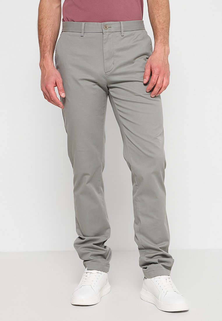 Hellegraue Chinos aus Baumwolle mit schlanker Passform, ausgestattet mit einem Knopfverschluss, Gürtelschlaufen und Taschen. Kombiniert mit weißen Sneakers.