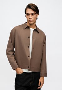 Άνδρας που φοράει καφέ σακάκι με κουμπιά πάνω από λευκό πουκάμισο και μαύρο παντελόνι, κοιτάζει προς τα αριστερά του με το ένα χέρι στην τσέπη του.