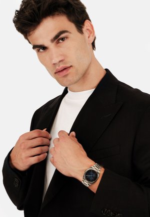 Sølvarmbandsur med en mørkeblå urskive, båret på en hånd der griber fat i en sort blazer. Hvid skjorte under, der viser et skræddersyet look.