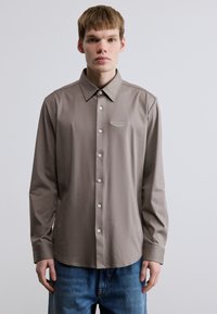 Καφέ μπλούζα με μακριά μανίκια και γιακά με κουμπιά, διακοσμημένη με μικρό κεντητό λογότυπο στο στήθος και λεία υφή υφάσματος.