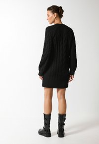 Abito in maglia nera a treccia con maniche lunghe, orlo a coste e motivo testurizzato. Indossato con stivali neri alti fino al ginocchio con dettaglio a fibbia.