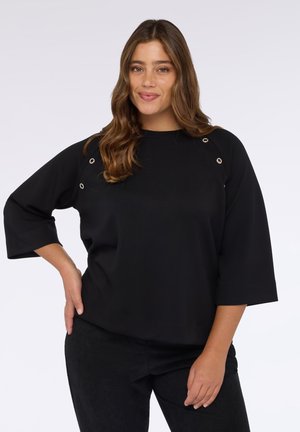 Maglietta nera con maniche 3/4, caratterizzata da occhielli argentati sulle spalle. Tessuto liscio con una vestibilità comoda e scollo rotondo. Abbinata a pantaloni neri.