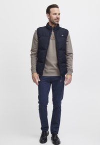 Marineblå puffervest over en grå hettegenser, parret med marineblå bukser og sorte sko. Vestene har glidelås foran og høy krage.