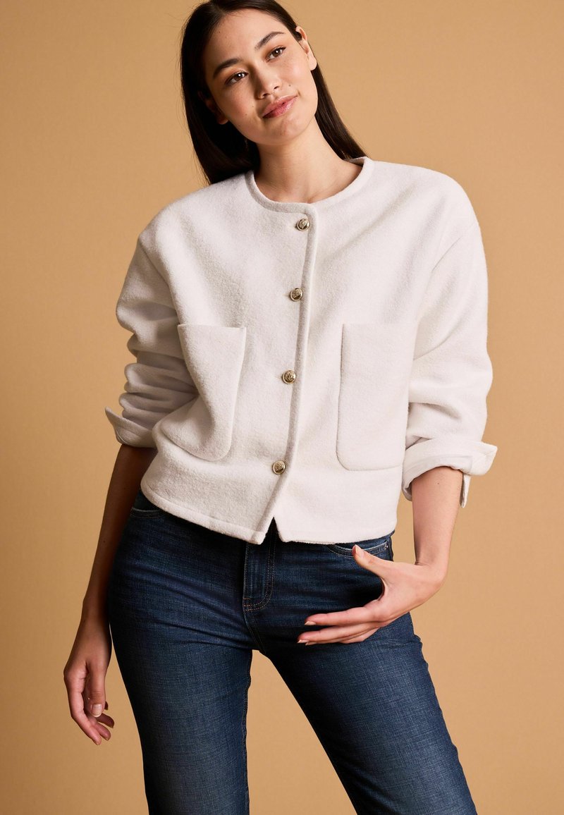 Weißer Fleece-Jacke mit Rundhalsausschnitt, vier Knöpfen und zwei vorderen Taschen. Kombiniert mit dunklen Jeans, vor einem neutralen Hintergrund.