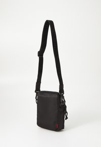 Borsa a tracolla nera in nylon testurizzato, dotata di chiusura a zip, tracolla regolabile e un piccolo tag rosso con logo.