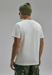 Biała bawełniana koszulka z krótkimi rękawami, okrągłym dekoltem i luźnym krojem, w połączeniu z bojówkami w kamuflażu oraz zieloną wełnianą czapką.