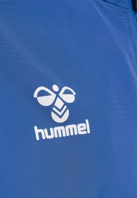 Blå sportstyg med en vit Hummel-logotyp som har en stiliserad design av två vingar och ett chevronmönster nedanför. Slät textur.