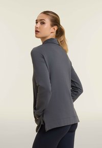 Maglia grigia a maniche lunghe con colletto, fessure laterali all'orlo e una texture liscia. Indossata con leggings scuri, mostrata da un angolo leggermente inclinato.