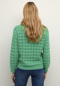 Pulover verde tricotat, cu un model texturat și tiv ribflat, asortat cu blugi albaștri. Designul include detalii lucrate în aer.