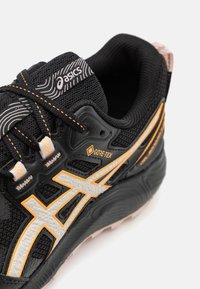 Scarpa atletica nera con branding Gore-Tex, caratterizzata da una tomaia testurizzata, dettagli arancioni e bianchi, e un design della suola imbottita.