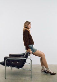 Brun pelskravejakke med en cropped design, parret med en denimnederdel. Model siddende på en sort lænestol med kromramme.