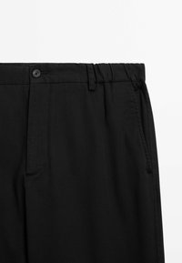 Black trousers in a smooth fabric with a relaxed fit, featuring a button closure, elastic waistband, and side pockets.