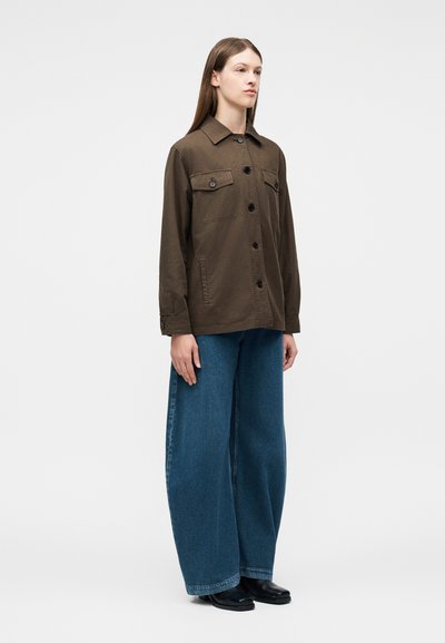 Γυναίκα στέκεται φορώντας καφέ μακριά ζακέτα με κουμπιά και τσέπες στο στήθος, με φαρδιά μπλε τζιν παντελόνια και μαύρα παπούτσια, σε απλό φόντο.