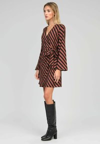 Femme portant une robe enveloppante à rayures diagonales marron et noires avec des manches longues et des bottes noires à talons hauts, se tenant devant un fond gris.