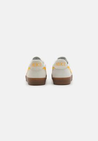 Nike tenisky s svěle šedým semišovým svrškem, oranžovými akcenty na boku a hnědou gumovou podrážkou. Obsahují logo značky na patě.