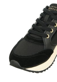 Black sneaker with mesh and suede materials, featuring gold eyelets, flat laces, and a white midsole with a textured black outsole.