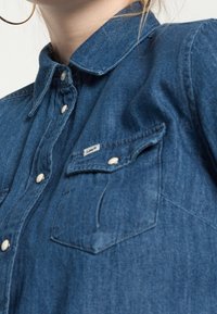 Denim shirt in dark blue, featuring a pointed collar, snap buttons, and a chest pocket with a branded label. Smooth texture and casual design.