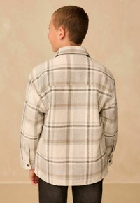 Camisa de franela a cuadros en colores crema, gris y marrón, con cuello, puños abotonados y corte relajado, que presenta un patrón a cuadros en toda su superficie.