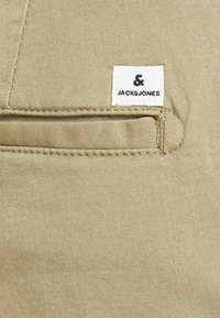 Beige bomullstyg med detalj av sidoficka. Har en vit etikett med svart text som säger "& JACK & JONES." Texturerad väv.