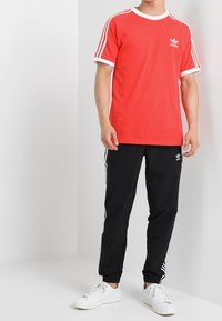 Punainen puuvillainen t-paita valkoisilla yksityiskohdilla ja Adidas-logo, yhdistettynä mustiin housuihin, joissa on valkoiset raidat, sekä valkoisiin lenkkareihin.