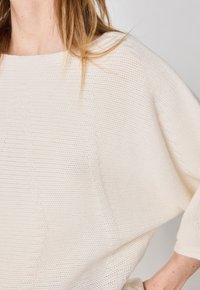 Pull en tricot texturé de couleur crème avec un décolleté large et une coupe ample, présentant des accents côtelés le long des coutures et des poignets.
