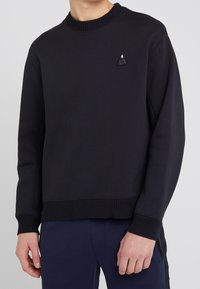 Sudadera negra de tela suave, con cuello redondo, puños y dobladillo acanalados, y un pequeño logo triangular en el pecho izquierdo.