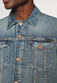 Jeansjacka i ljusblått med en blekt textur, med kopparfärgade knappar, två bröstfickor och en klassisk krage.