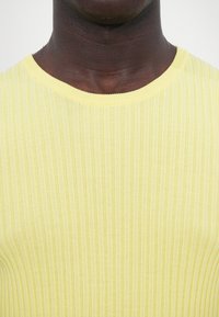 Pulover tricotat cu răsuciri galbene, având un guler rotund și un material texturat, iar imaginea de aproape evidențiază detaliile de culoare și model.