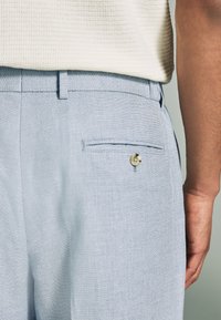 Helle blaue Hose aus strukturiertem Stoff, mit seitlichen Taschen und einer Gesäßtasche mit Knopfdetail, seitlich aufgenommen.