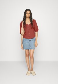 Blusa de botão cor ferrugem com mangas balão, combinada com uma saia mini de ganga azul claro e sapatos slip-on bege. Modelo segura uma pequena mala.