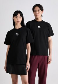Čierne bavlnené tričká s krátkymi rukávmi a okrúhlym výstrihom, s bielym logom Lacoste na hrudi, nosené s koženou sukňou a bordovými nohavicami.