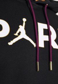 Sweat à capuche noir avec un logo Jordan doré et un texte blanc. Il présente des cordons de serrage violets avec des embouts métalliques dorés et un tissu texturé.