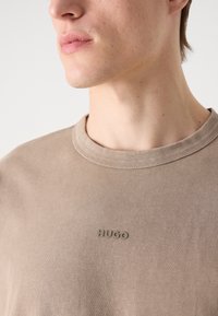 T-shirt en coton marron avec un col rond, arborant un logo "HUGO" discret dans une teinte assortie sur la poitrine. Texture douce, design décontracté.