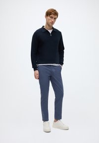 Πλεκτή μπλούζα με φερμουάρ σε ναυτικό μπλε χρώμα και γιακά, συνδυασμένη με ανοιχτό μπλε παντελόνι και λευκά αθλητικά παπούτσια. Ομαλή υφή, μοντέρνα εφαρμογή και λιτός σχεδιασμός.