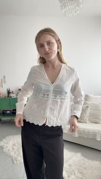 Jeune femme aux cheveux blonds portant un chemisier en dentelle blanche et un pantalon noir, debout dans un salon lumineux avec un canapé beige et une armoire verte.