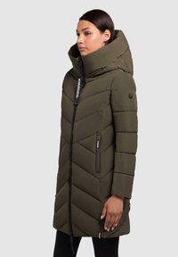 Olivgrön puffjacka med hög krage, sicksack-quilting, framdragkedja och sidofickor. Har ett svart logotag på dragkedjan.