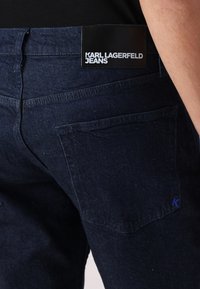 Sötétkék farmernadrág sima felülettel, fekete bőr márkajelzéssel és egy kis, lila hímzett 'K'-val a hátsó zsebben.