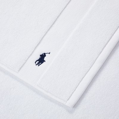 Λευκή πετσέτα από πετσέτα βελουτέ με κεντητό λογότυπο παίκτη πόλο σε ναυτικό μπλε. Η υφή είναι μαλακή με λεπτομέρεια ριγωτής άκρης.