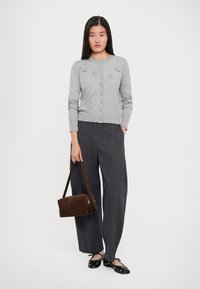Donna che indossa un cardigan grigio con bottoni e dettagli floreali, pantaloni larghi neri, ballerine nere, con una borsa marrone a lato.