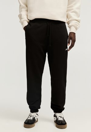 Mężczyzna ubrany w czarne spodnie dresowe ze ściągaczem, białe sneakersy z czarnymi detalami oraz kremową bluzę, stojący na neutralnym tle.