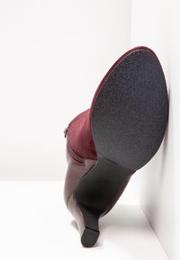 Botte à talon bordeaux pour femme avec semelle noire texturée, présentée reposant sur son talon contre un fond blanc en coin.