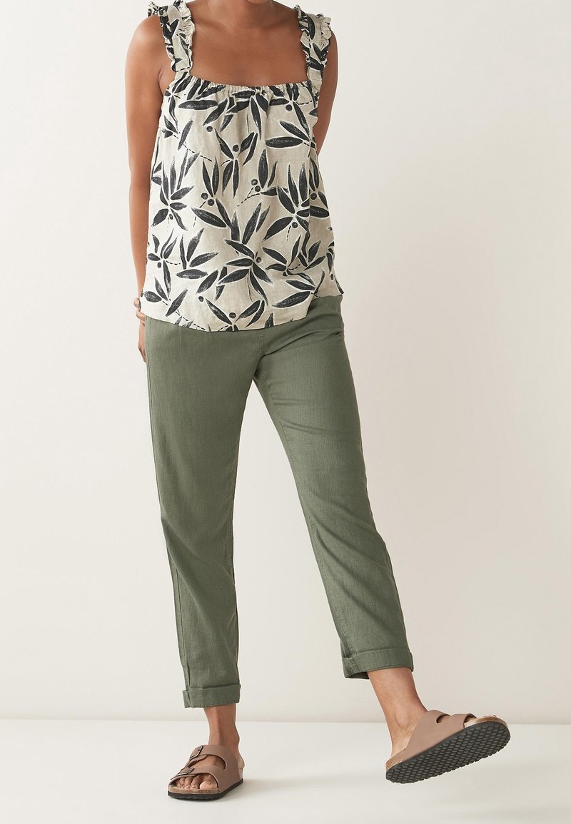 Personne portant un haut blanc sans manches avec un motif de feuilles noires, un pantalon vert olive roulé aux chevilles, et des sandales marron à enfiler.