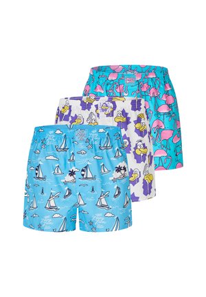 Muskelbepackter Mann mit kurzen geflochtenen Haaren trägt hellblaue Badeshorts mit weißen und dunkelblauen Segelboot- und Wolkenmustern, der vor einem schlichten Hintergrund steht.